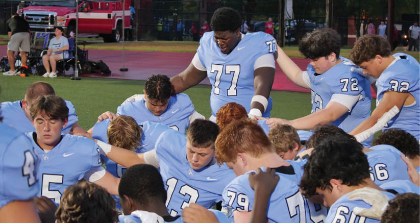 A group of people wearing light blue sports jerseys are gathered closely on a field, some with heads bowed and one person standing with arms resting on others' shoulders. Description generated by AI
