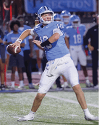 A person wearing a light blue football uniform with the number 10 is preparing to throw a football on a field with other players and coaches in the background. Description generated by AI