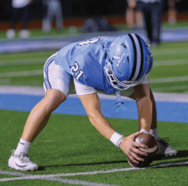 A person in a blue football uniform is bending down on a field to place a football on the ground. Description generated by AI