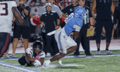 A person in a blue football uniform is running while another person in a white uniform is on the ground reaching out to tackle them during a nighttime football game. Description generated by AI