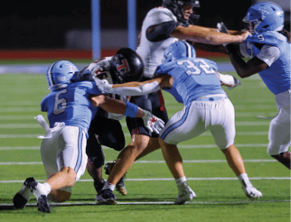 Two people in light blue football uniforms are tackling a person in a black and white football uniform on a green field during a game. Description generated by AI