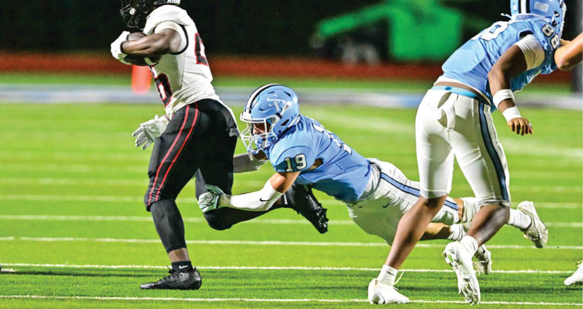 A person in a light blue football uniform is diving to tackle a person in a black and white football uniform who is running with the ball on a green field. Description generated by AI
