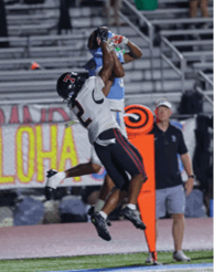 Two people in football uniforms jump to catch a football near the sideline while two people stand in the background watching. Description generated by AI