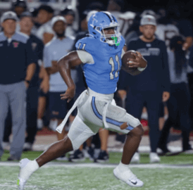 A person in a blue football uniform is running with the ball on a field while another person in a black and white uniform is approaching from behind. Description generated by AI