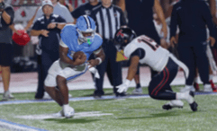 A person in a light blue football uniform is running with the ball while another person in a white and black uniform is crouching and preparing to tackle on a football field. Description generated by AI