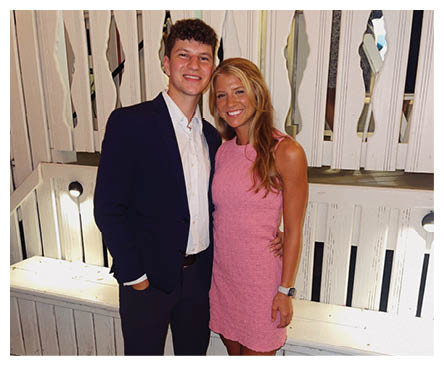 Two adults stand close together with one wearing a pink sleeveless dress and the other in a dark suit with a white shirt, against a white wooden background. Description generated by AI