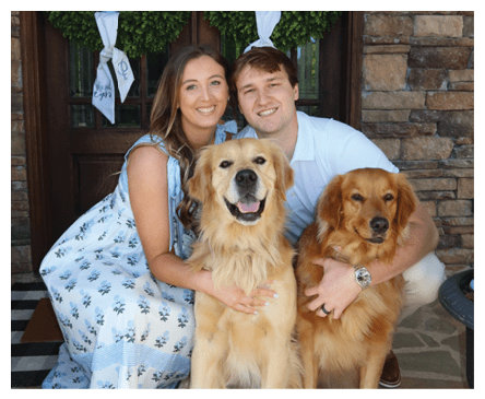 Two adults smiling and sitting closely together on a porch with two golden retrievers, one wearing a watch and ring, with wreaths hanging on the door behind them. Description generated by AI
