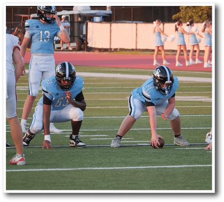 People in football uniforms are positioned on a field preparing for a play while cheerleaders stand in the background. Description generated by AI