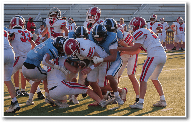 A group of people wearing football uniforms are engaged in a tackle on a sunlit field with empty bleachers in the background. Description generated by AI