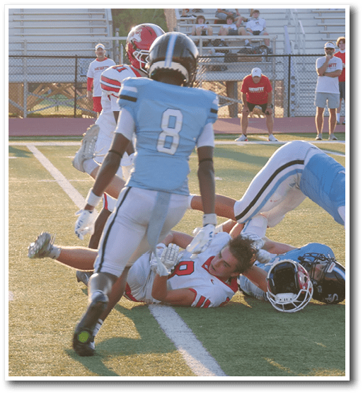 A group of people playing football on a field with one person in a light blue uniform standing over another person in a red and white uniform lying on the ground holding the ball. Description generated by AI
