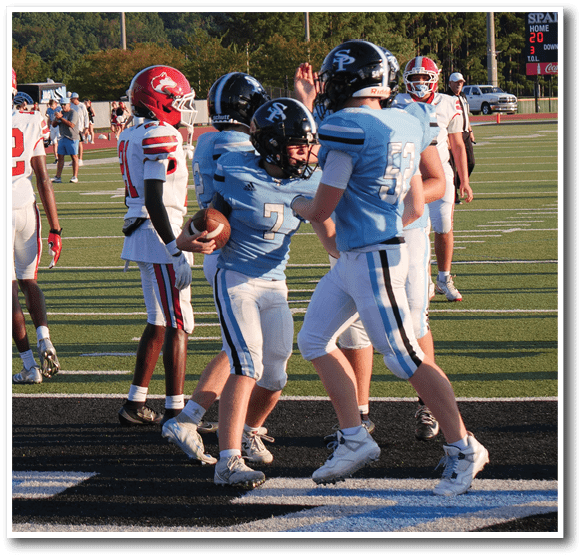 People wearing football uniforms are on a field with some players in light blue and others in white and red, with one player holding a football. Description generated by AI
