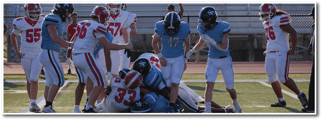 People wearing football uniforms are engaged in a tackle on a grassy field during a game. Description generated by AI