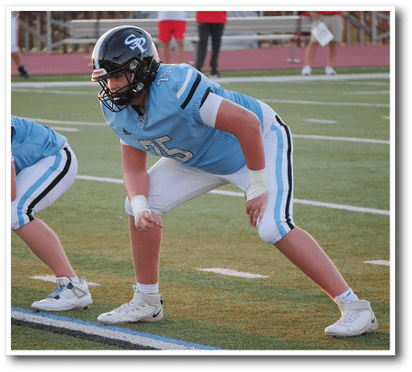 A person in a light blue football uniform and black helmet is crouched in a ready position on a football field. Description generated by AI