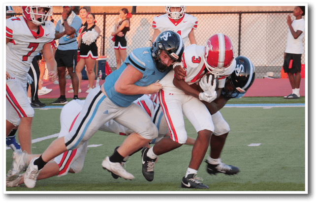 Two people in football uniforms are tackling a third person holding a football on a field while others and cheerleaders watch in the background. Description generated by AI