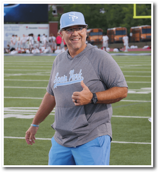 A person wearing a gray Spain Park shirt, blue shorts, and a light blue cap is smiling and giving a thumbs-up on a sports field with blurred people and school buses in the background. Description generated by AI