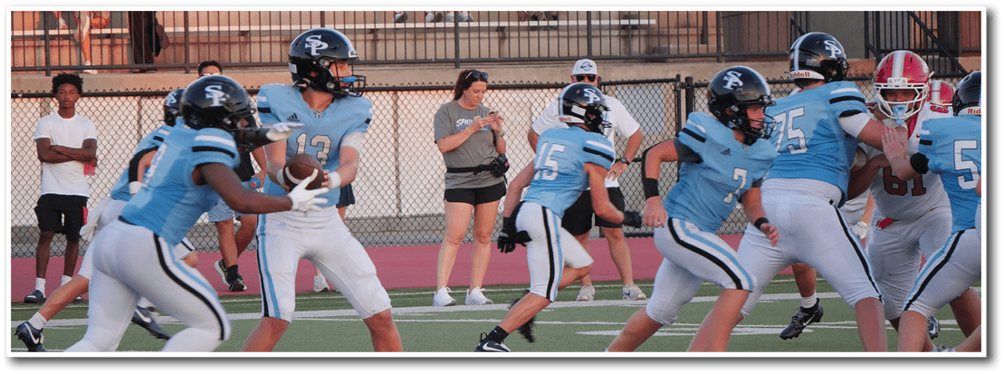 A group of people in football uniforms are on a field with one person holding a football while others prepare to block or run, and several people are watching from the bleachers in the background. Description generated by AI