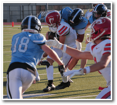 People wearing football helmets and uniforms are engaged in a play on a football field with one person holding the ball tightly while others attempt to tackle. Description generated by AI