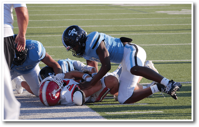 Three people in football uniforms are on a field with two players in light blue and white tackling a player in a red and white uniform who is lying on the ground. Description generated by AI