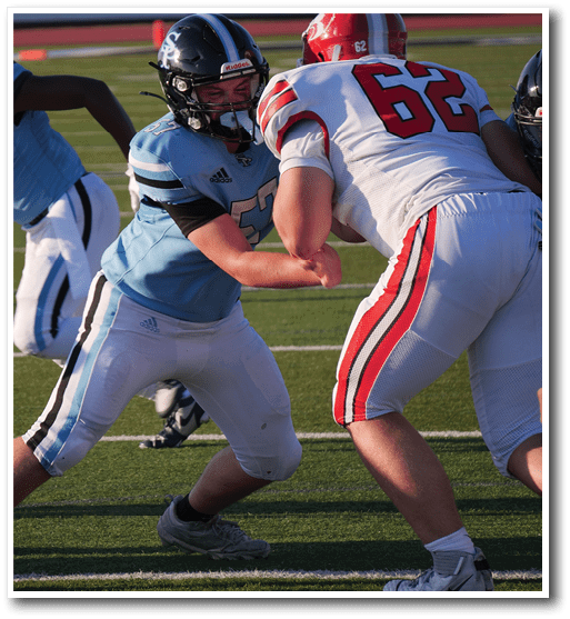 Two people in football uniforms and helmets are engaged in a blocking stance on a grassy field during a game. Description generated by AI