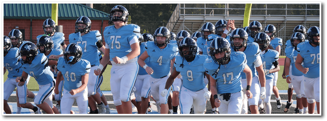 People wearing light blue football jerseys and white pants are running onto a field from a track area. Description generated by AI