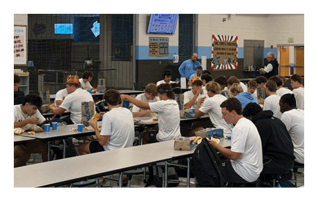 People wearing white shirts are sitting at cafeteria tables with attached stools, eating and interacting. Description generated by AI
