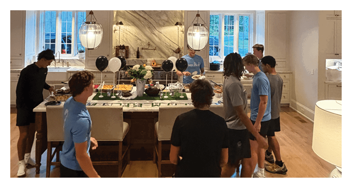 A group of people wearing casual clothing are gathered around a kitchen island in a warmly lit, rustic-style kitchen with wooden beams and large windows. Description generated by AI
