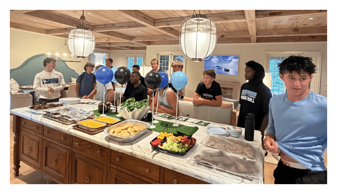Several people are gathered around a table with food in a warmly lit room with wooden ceiling beams and cabinets. Description generated by AI