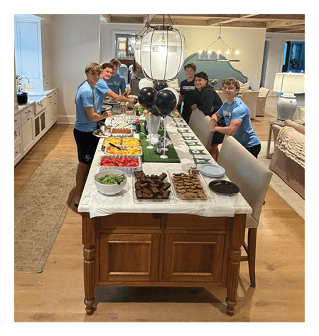 A group of six people wearing casual blue and black shirts are gathered around a long table set with various foods and black and white balloons in a warmly lit kitchen and dining area. Description generated by AI