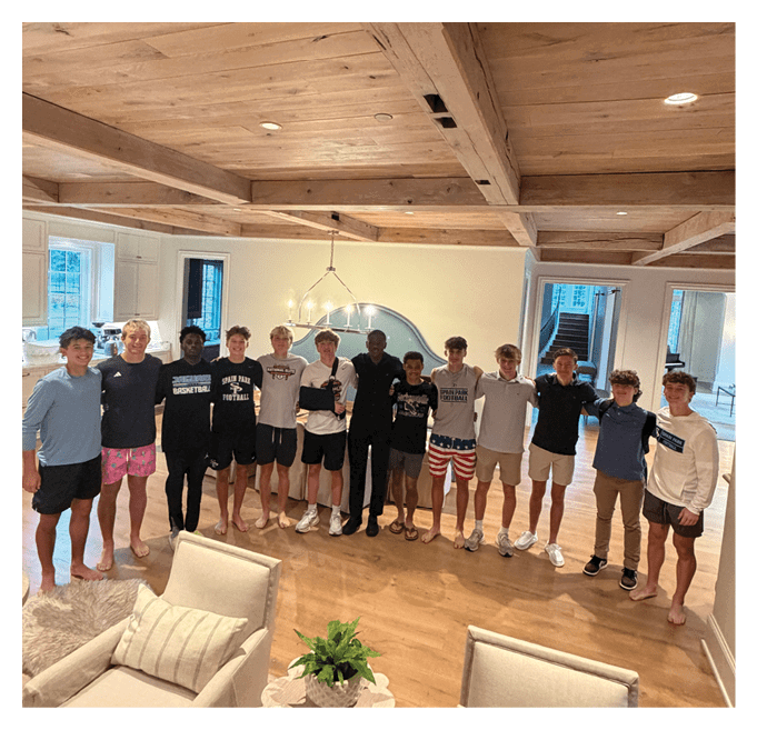 A group of people standing closely together in a well-lit room with wooden beams on the ceiling and casual clothing, some barefoot and some wearing shoes. Description generated by AI