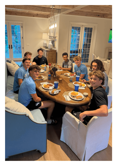 A group of eight people sitting around a dining table enjoying a meal with plates of food and blue cups in a warmly lit room. Description generated by AI