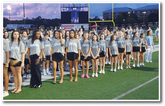 A group of people wearing matching gray shirts and standing in a line on a sports field during an evening event with a scoreboard and goalposts in the background. Description generated by AI