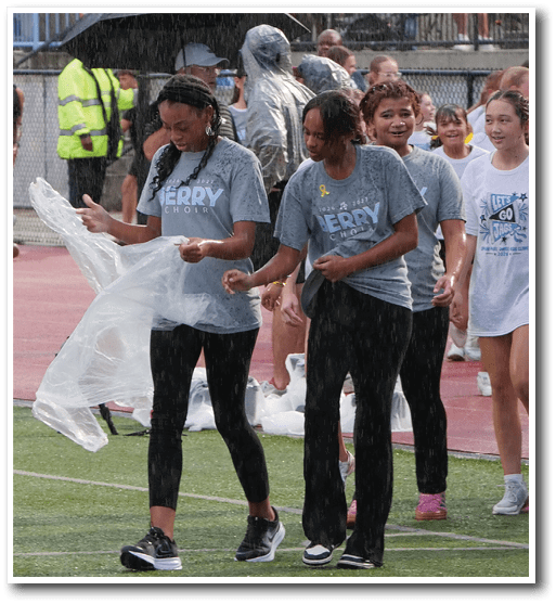 People wearing matching event t-shirts walk on a rainy sports field while others watch from bleachers in the background. Description generated by AI