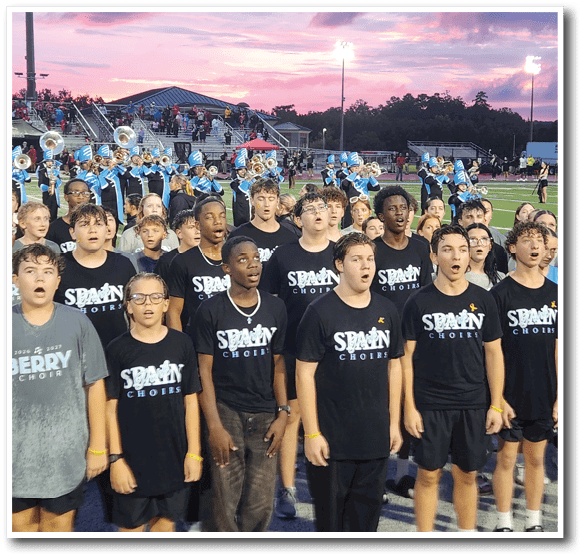 A group of people wearing matching black shirts with “Spain Choirs" printed on them are standing and singing on a sports field with a marching band playing instruments in the background during sunset. Description generated by AI