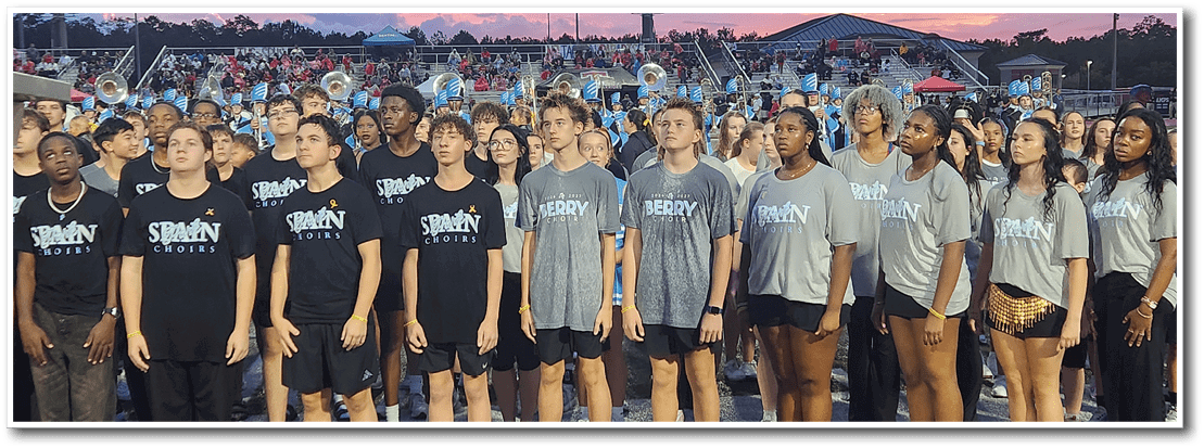 A group of people wearing matching black and gray shirts stand in a line on a field with a crowd and band visible in the background. Description generated by AI