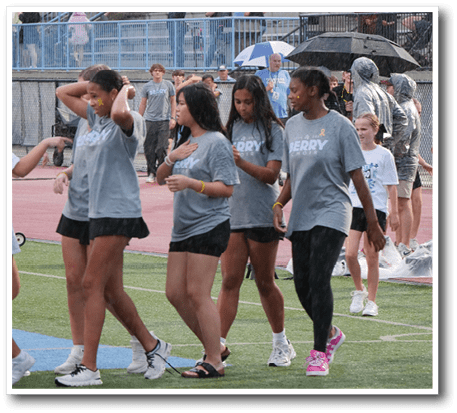 A group of people wearing matching gray shirts and black shorts or leggings are walking on a sports field with some spectators and umbrellas visible in the background. Description generated by AI