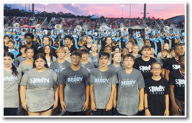 A large group of people wearing matching choir t-shirts stand in front of a marching band performing at an outdoor event during sunset. Description generated by AI