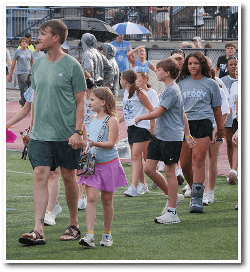 A group of people, including children and adults, are walking on a sports field with some wearing casual athletic clothing and one person wearing a medical walking boot. Description generated by AI