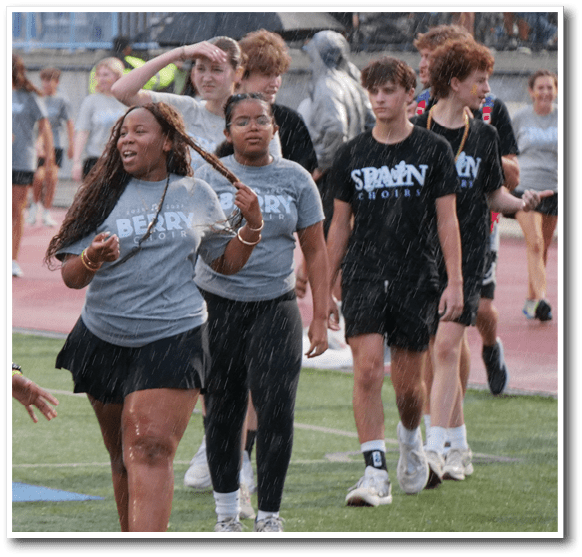 A group of people wearing matching gray shirts and walking on a sports field in the rain. Description generated by AI
