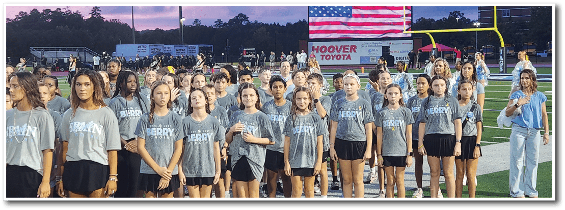 A group of people wearing matching gray shirts and black shorts stand in rows on a sports field with a banner and goalposts in the background during dusk. Description generated by AI