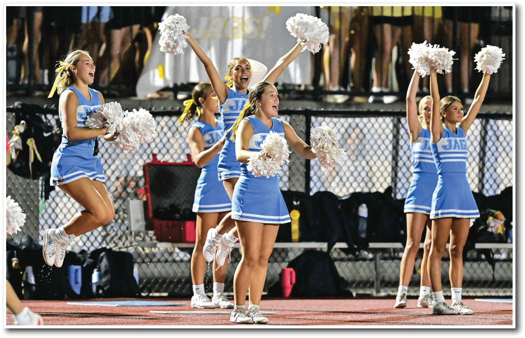 People wearing athletic shoes and cheerleading uniforms stand on a sports field near a running track. Description generated by AI