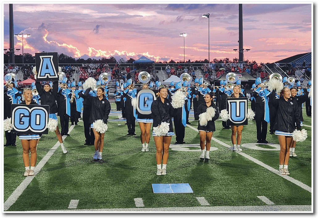 A group of adults in cheerleading uniforms and jackets stand on a football field holding pom-poms and signs that spell out “GO CU" with a marching band playing in the background. Description generated by AI