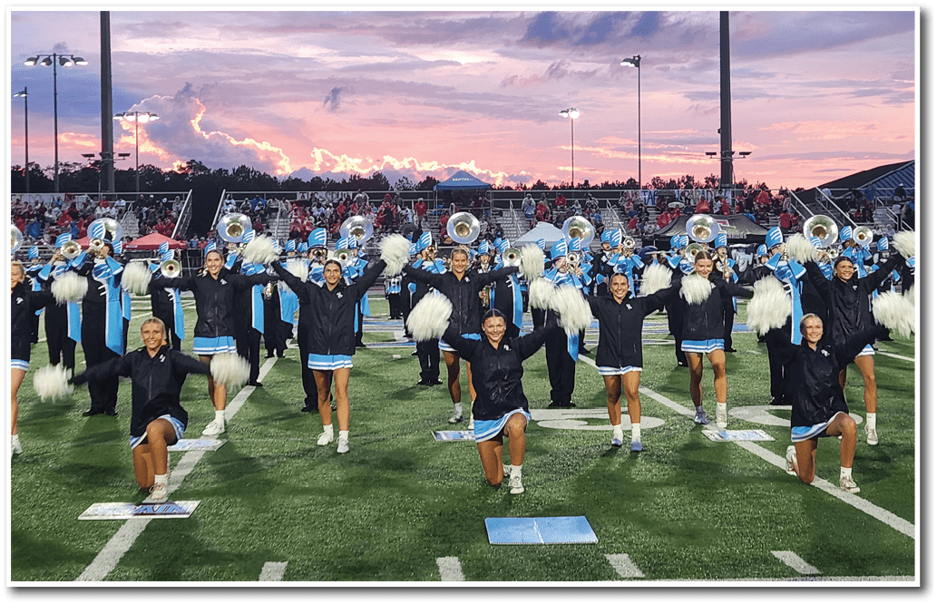 A group of people in matching black jackets and blue skirts are performing a coordinated cheerleading or marching band routine on a football field at sunset. Description generated by AI