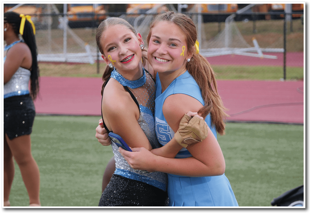Two smiling people in cheerleading outfits embrace on a sports field with a track and goalposts in the background. Description generated by AI