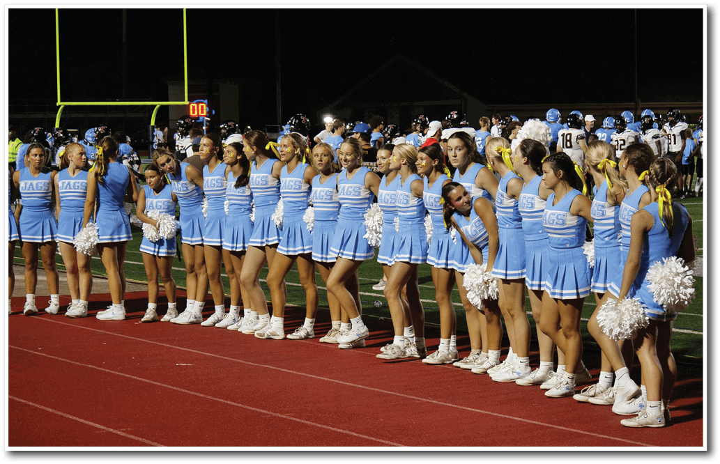 A group of adults in blue cheerleading uniforms stand in a line on a track at night near a football field with players and goalposts visible in the background. Description generated by AI