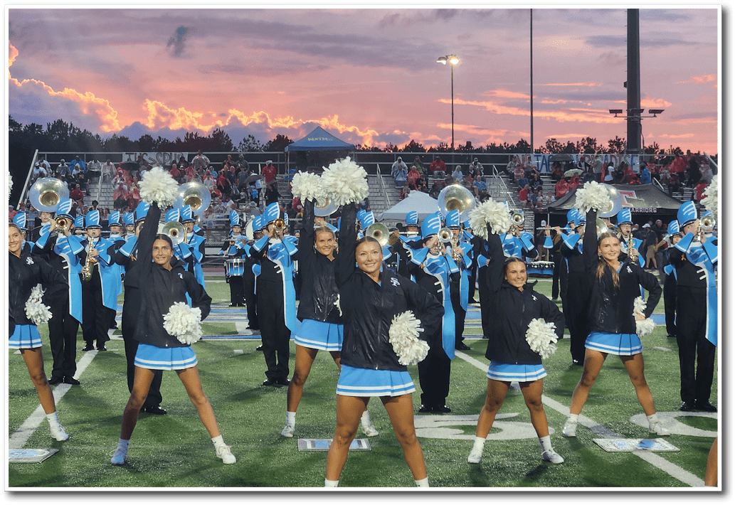 People in cheerleading uniforms and jackets perform with pom-poms on a football field while a marching band plays behind them during sunset. Description generated by AI