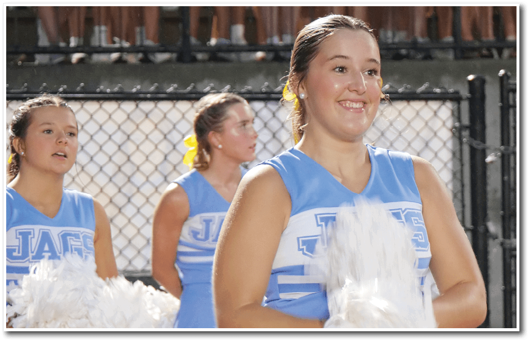 A person in a blue cheerleading uniform is smiling and holding white pom-poms with other cheerleaders and a chain-link fence in the background. Description generated by AI