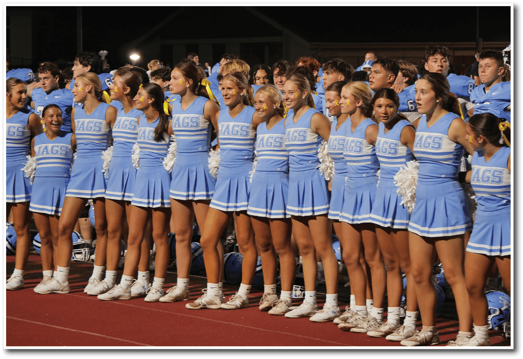 A group of adults in matching blue cheerleading uniforms with “JAGS" on the front stand side by side on a track, some holding white pom-poms, with a crowd and players in the background. Description generated by AI