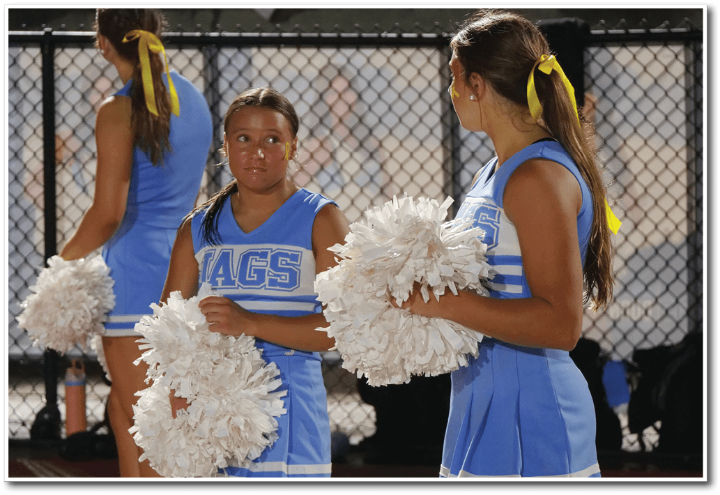 Three people in blue cheerleading uniforms with yellow hair ribbons hold white pom-poms while standing near a chain-link fence. Description generated by AI