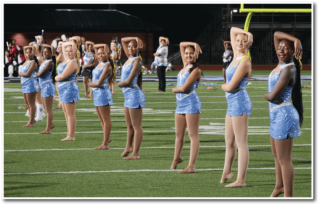 A group of people dressed in sparkly blue dance costumes are standing in a line on a football field performing a synchronized pose with one arm bent over their heads. Description generated by AI
