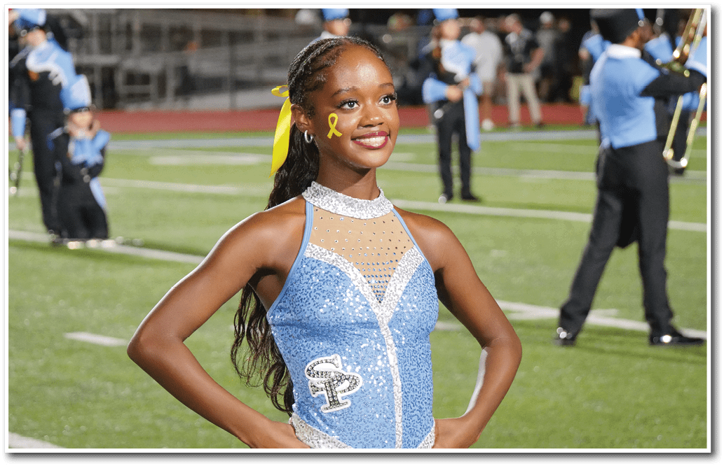 A person wearing a sparkly blue and white costume with a yellow ribbon on their face and hair stands smiling on a sports field with band members in the background. Description generated by AI